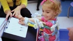 Emma usando exoesqueleto impresso em 3D. Criança com giz de cera na mão, pintando desenho em cima de uma mesa. Emma está usando blusinha rosa com listras brancas. Exoesqueleto customizado com desenhos em todo o redor.
