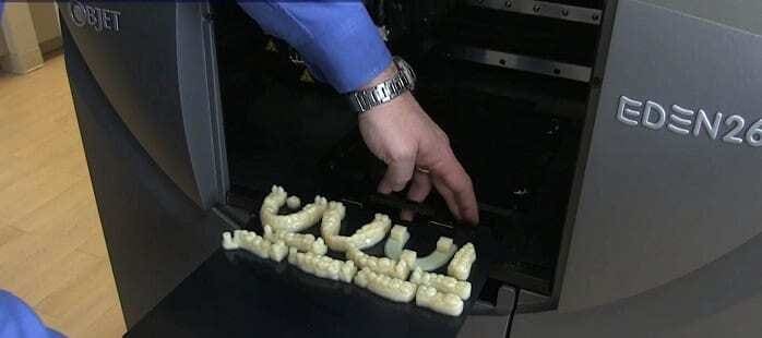 Modelos de Próteses dentárias saindo de impressora 3D. Mão puxando as próteses de dentro da impressora. Continue lendo nosso post.