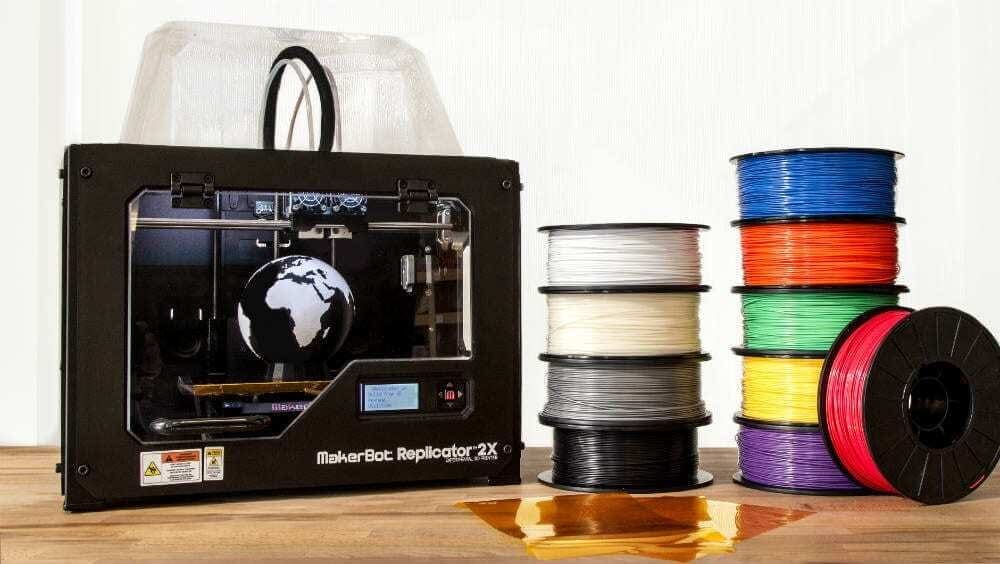 Rolos para serem usado em impressora 3D amontuados, um em cima do outro com cores diferentes (Branca, bege, cinza, preta, azul. vermelho, verde, amarelo e roxo) e impressora Makerbot Mini ao lado, com o globo terrestre impresso dentro.